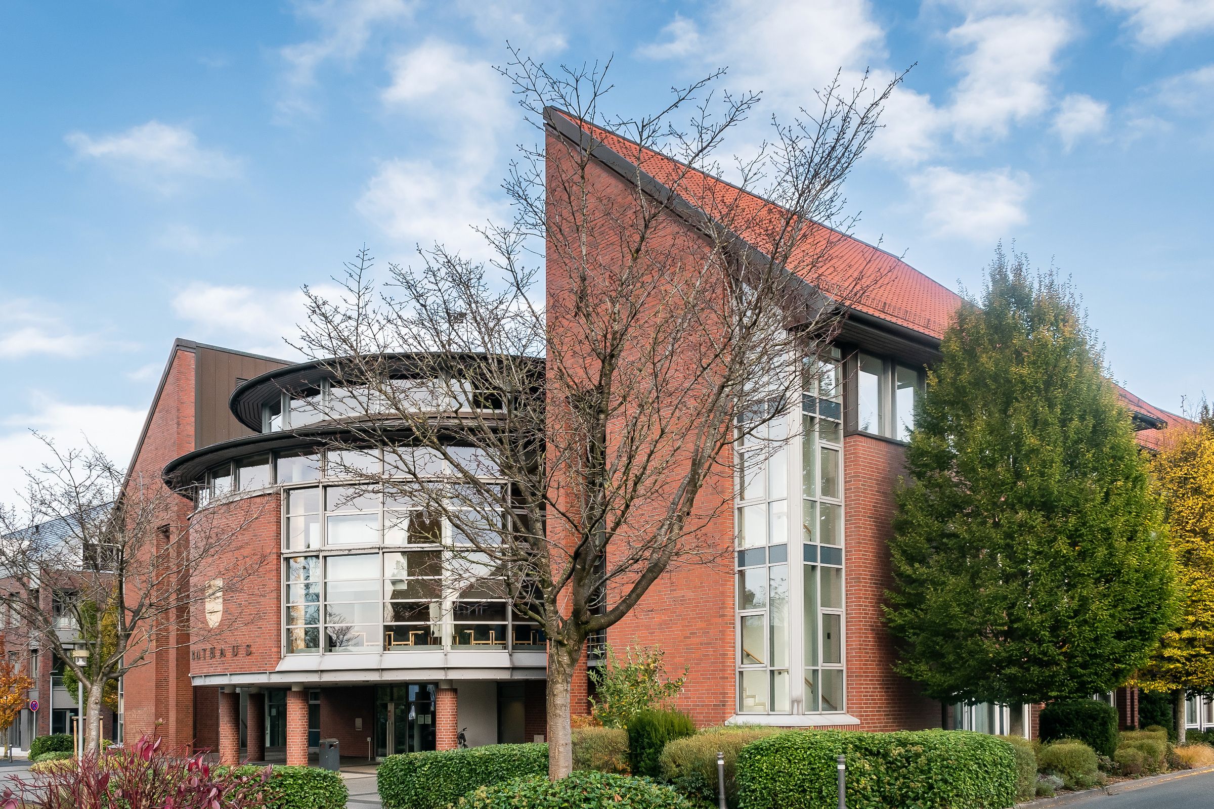 Das Rathaus in Cloppenburg mit geschwungener Glasfassade zwischen Bäumen und Hecken.