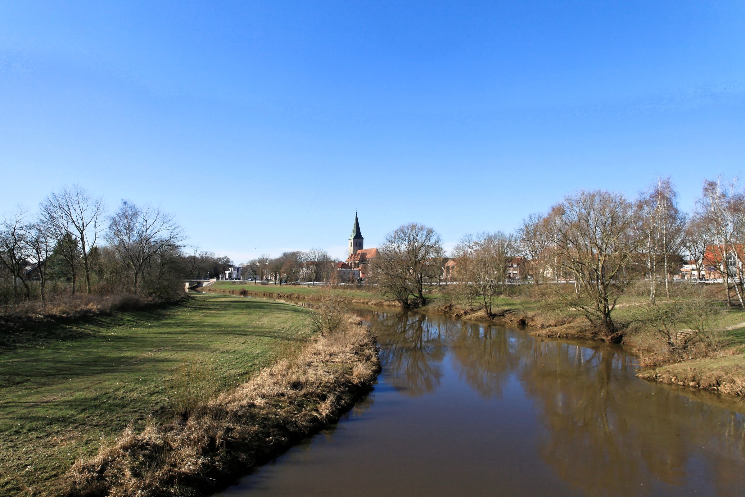 Die Hase in Haselünne mit grünen Ufern und der Kirche St. Vincentius im Hintergrund.